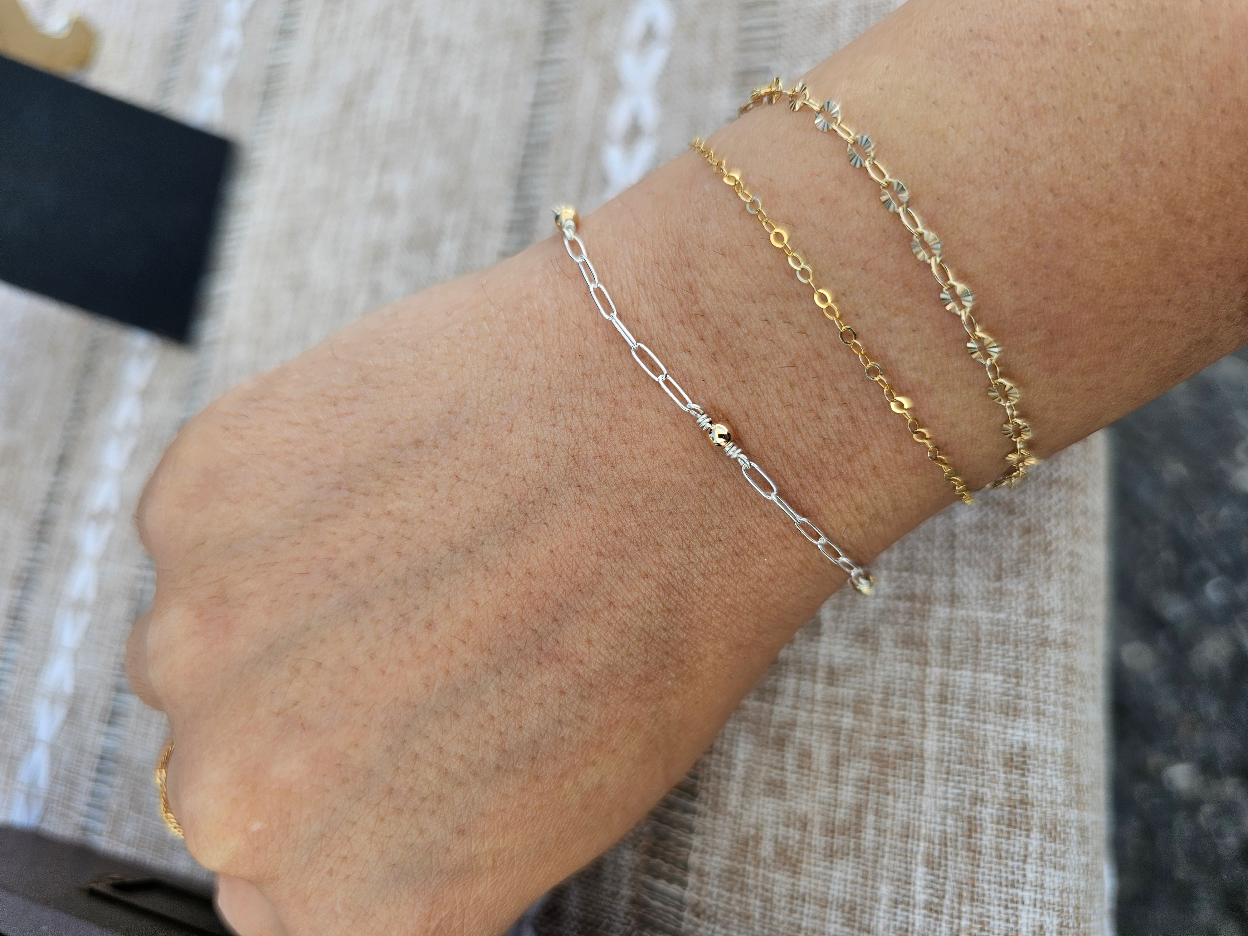 Close-up of a wrist with three stacked delicate bracelets — two thin gold chains (one with tiny floral links) and a silver paperclip chain with a small gold bead, photographed on a beige woven tablecloth.