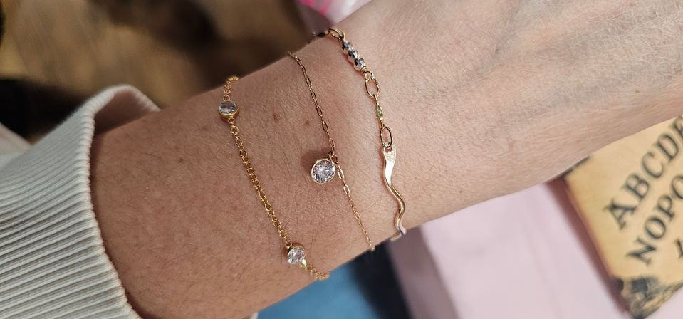 Close-up of a wrist wearing three stacked delicate gold bracelets — two fine chains with sparkling bezel-set clear stones and one wavy gold link, pictured against a ribbed cream sweater cuff and blurred book background.