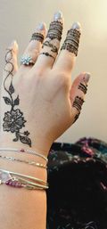 Close-up of a hand with black henna (mehndi) floral and finger-band designs, silver stacked bracelets, a sparkly cluster ring, and metallic-painted nails.