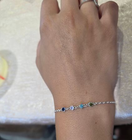 Close-up of a hand wearing a delicate silver chain bracelet with four small round gemstones—navy blue, light purple, aqua blue and olive green—paired with a thin silver ring, photographed on a cream fabric background.