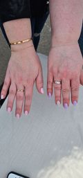 Close-up of two hands on a sunlit outdoor table showcasing a pastel manicure: left with glossy pale-pink nails, gold rings and bracelets; right with lavender and confetti-speckled nails and a silver ring.