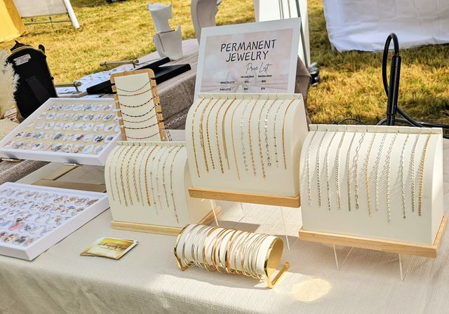 Sunlit outdoor artisan market jewelry display featuring a 'Permanent Jewelry' sign and rows of delicate gold and silver bracelets and anklets on white stands at a vendor booth.