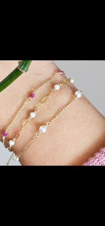 Close-up of a wrist wearing layered delicate gold chain bracelets with small freshwater pearls, textured matte gold beads and pink gemstone accents, minimalist stacked bracelet jewelry.