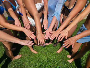 Hands of a group of friends in swimsuits reaching together over a green lawn at a sunny poolside gathering, showing bracelets, rings and colorful manicures.