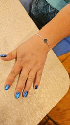 Close-up of a hand resting on a beige textured table, showcasing metallic blue glitter nails and a delicate silver chain bracelet with a round dark stone.