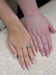 Two hands side-by-side on a pale satin surface, one with almond-shaped white nail art featuring stars and bows and stacked rings, the other with short natural nails; both wear delicate silver bow bracelets.