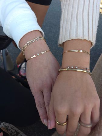 Close-up outdoor shot of two hands wearing delicate gold bracelets and a chain-link bangle with stacked rings and knit sleeves — casual street-style jewelry