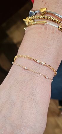 Close-up of wrist with stacked gold and silver bracelets — delicate gold chains with tiny beads and pearls, a beaded gold strand, and a silver cable bangle with heart and cross charms on a blurred background.