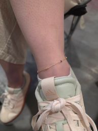 Close-up of an ankle wearing a delicate gold chain anklet with a small pink bead, resting on a mint-and-cream pastel sneaker with knotted laces — casual street style.