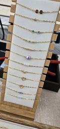 Row of delicate gold chain bracelets with tiny gemstone accents, pearls, beads and heart and disc charms neatly arranged on a white leather panel set in a bamboo wooden display stand — stylish tabletop jewelry display.
