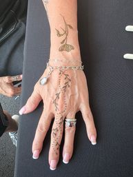 Close-up of a hand with delicate henna vine and dotted floral design running from wrist to middle finger, wearing silver chain bracelets, a gemstone ring and French-manicured nails on a black table — artisan henna tattoo and hand jewelry close-up.