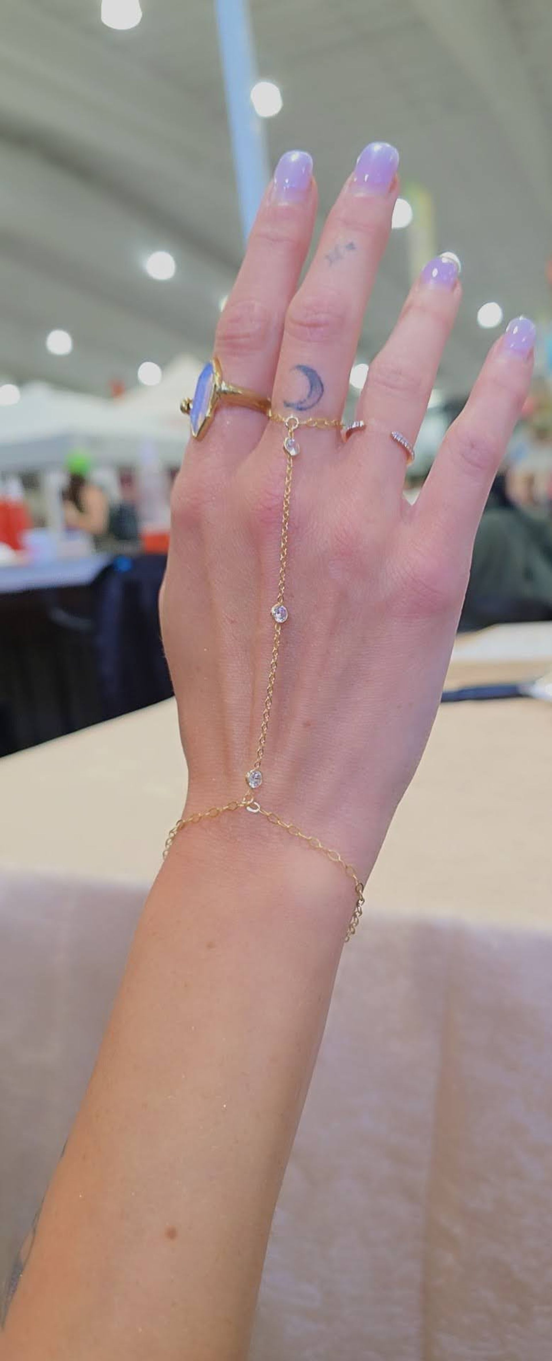 Close-up of a hand with lavender ombre nails and a crescent moon finger tattoo wearing a gold hand-chain that links a statement ring to a delicate bracelet with small clear stones, blurred indoor event background.