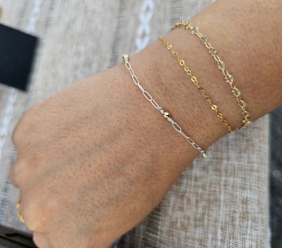 Close-up of a wrist with three stacked delicate bracelets — two thin gold chains (one with tiny floral links) and a silver paperclip chain with a small gold bead, photographed on a beige woven tablecloth.