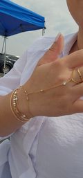 Close-up of hand in a white blouse wearing layered gold bracelets and a delicate gold hand-chain with crystal and pearl beads, photographed outdoors under a blue canopy.