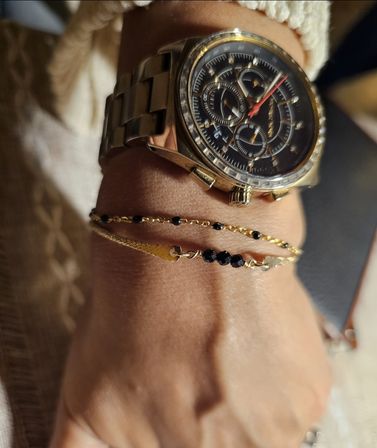 Close-up of a gold metal chronograph watch with black dial and red second hand paired with two delicate gold bracelets threaded with small black beads on a wrist