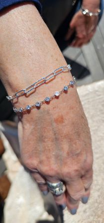 Close-up of a sunlit wrist and hand wearing two silver bracelets — a textured paperclip chain and a bezel-set clear-stone bracelet — paired with a hammered silver ring and gray nail polish.