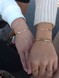 Close-up outdoor shot of two hands wearing delicate gold bracelets and a chain-link bangle with stacked rings and knit sleeves — casual street-style jewelry