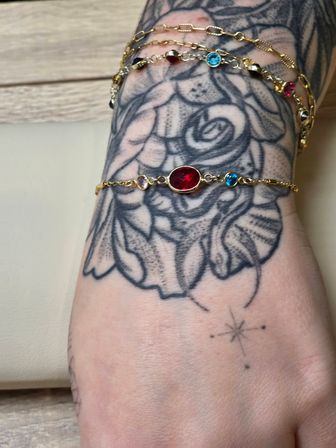 Tattooed wrist with black floral design wearing delicate gold chain bracelets centered on an oval red gemstone with small blue and pink stones and a tiny star tattoo on the hand
