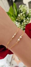 Close-up of a wrist wearing two delicate gold chain bracelets with alternating tiny gold beads and white pearls, photographed over a red rose and white floral background — minimalist fashion jewelry close-up.