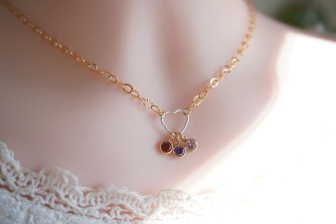 Close-up product photo of a dainty gold chain necklace with an open heart pendant and three small round red, purple, and pink gemstone charms, displayed on a pale mannequin bust with lace trim.