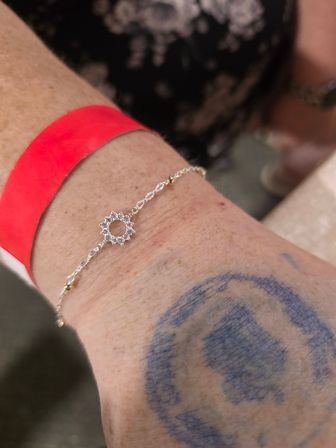 Close-up of a wrist wearing a delicate silver sunburst charm bracelet with tiny crystals, a bright red event wristband, and a faded blue circular ink stamp/tattoo.