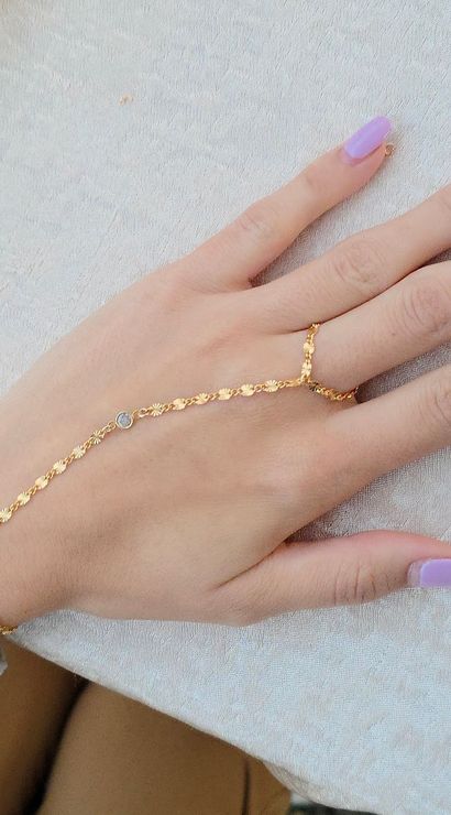 Close-up of a hand wearing a delicate gold hand chain connecting a bracelet to a ring, pastel lavender manicure, resting on a textured cream tablecloth.