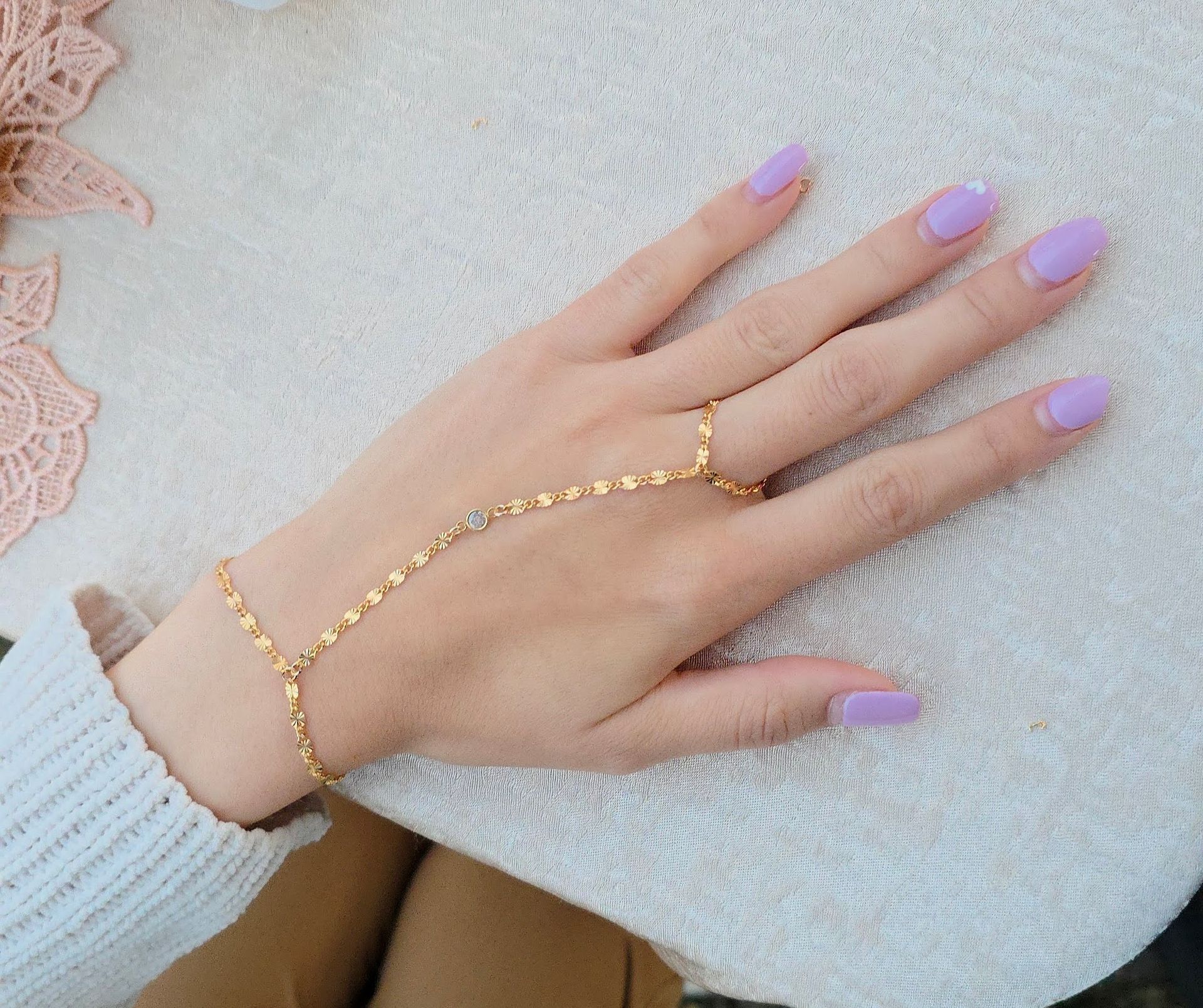 Close-up of a hand wearing a delicate gold hand chain connecting a bracelet to a ring, pastel lavender manicure, resting on a textured cream tablecloth.