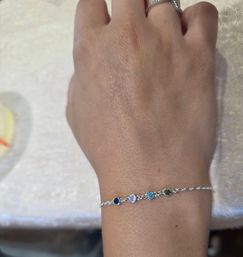 Close-up of a hand wearing a delicate silver chain bracelet with four small round gemstones—navy blue, light purple, aqua blue and olive green—paired with a thin silver ring, photographed on a cream fabric background.