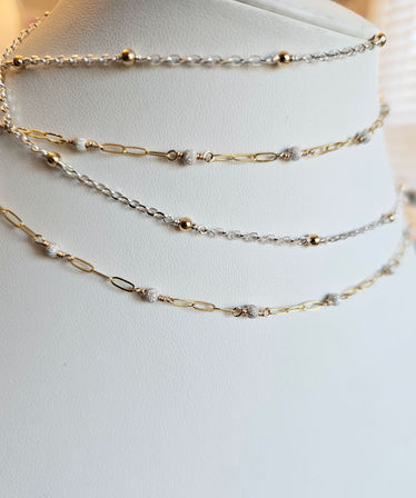 Stacked delicate gold and silver chain necklaces with tiny textured bead accents draped on a white leather jewelry display