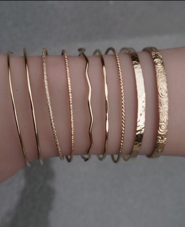 Close-up of a wrist wearing a stacked set of slim gold bangles with mixed textures — smooth, twisted, wavy, hammered and floral-engraved — minimalist boho jewelry accessory.