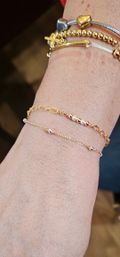 Close-up of wrist with stacked gold and silver bracelets — delicate gold chains with tiny beads and pearls, a beaded gold strand, and a silver cable bangle with heart and cross charms on a blurred background.