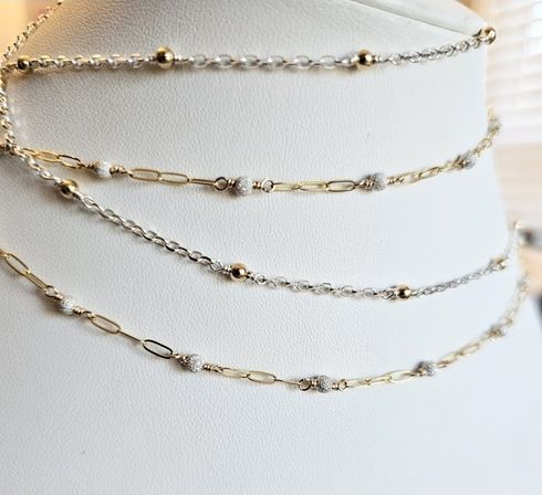 Close-up of delicate mixed-metal gold and silver chain necklaces with tiny textured and smooth bead accents, layered on a white jewelry display bust — fashion accessory photo.