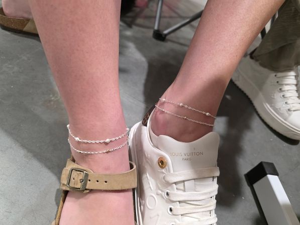 Close-up of two ankles wearing delicate double-chain pearl anklets — one foot in a tan suede buckle sandal, the other in a white designer-style sneaker on a concrete store floor.