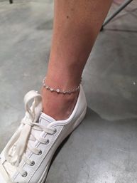 Close-up of an ankle wearing a delicate silver crystal-studded anklet paired with a white canvas sneaker on a concrete floor, casual summer accessory shot