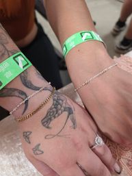Close-up of two wrists side-by-side wearing green event wristbands, delicate gold and silver chain bracelets, a tattooed hand with small bird and abstract tattoos, and a sparkling diamond engagement ring.