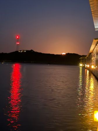 Dusk coastal harbor with red navigation tower lights reflecting on calm water and a glowing moonrise behind silhouetted hills, lights along a waterfront pier.