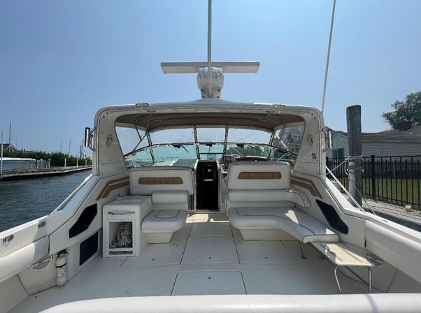 Spacious white motor yacht cockpit with cushioned wraparound seating, helm under a sunshade and clear windshield, docked at a sunny marina with pilings and calm water.