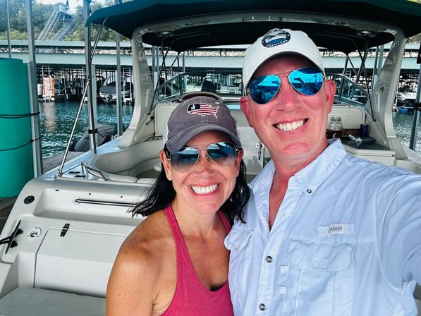 Smiling couple in sunglasses and baseball caps taking a selfie aboard a white motorboat at a covered marina dock with boats and calm water in the background