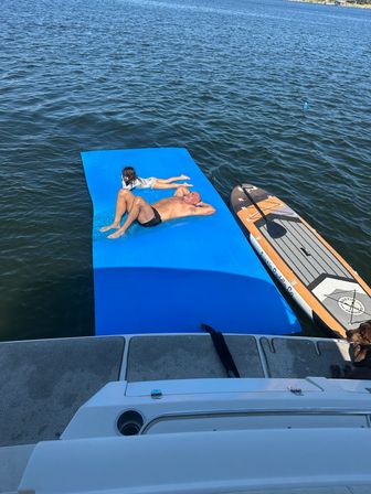Adult and child lounging on a bright blue floating mat beside a paddleboard at the stern of a boat on a sunny lake, dog peeking from the swim platform.