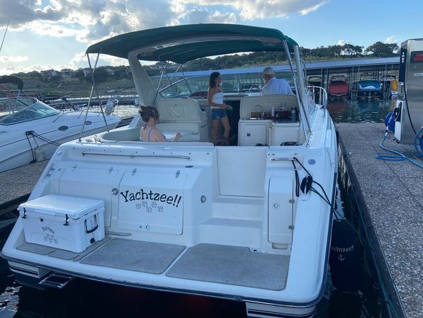 White motorboat named Yachtzee!! docked at a marina, passengers relaxing under a green bimini top, cooler on the swim platform, calm lake water and covered boat slips in the background.