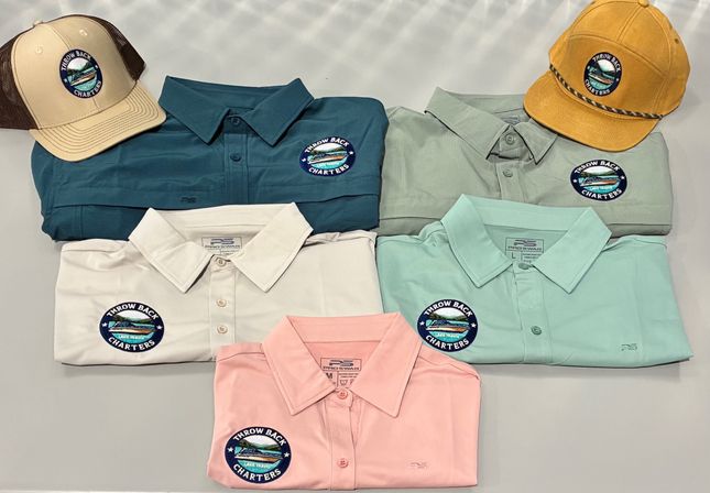 Stacked folded collared performance fishing shirts in pastel and neutral shades with two caps, each bearing a round embroidered nautical patch depicting a boat and waves — coastal outdoor apparel display.