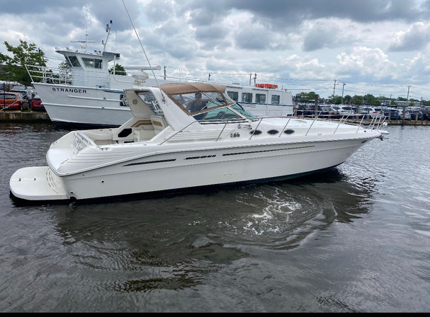 Sleek white cabin-cruiser motorboat gliding past a busy marina waterfront with docked vessels and parked cars under an overcast sky
