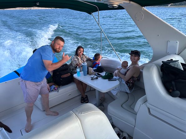 Group of friends and family on a motorboat enjoying a boat ride on open water, seated around a small table—one holding a baby and another giving a thumbs-up, boat wake and shoreline visible