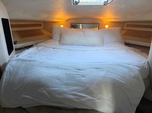 Cozy boat cabin v-berth with white linens, textured lumbar pillow, overhead hatch and warm reading lights — yacht interior sleeping area.
