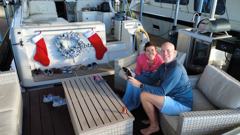 Couple relaxing on wicker sofa at a yacht's aft deck in a marina, cozy holiday wreath and red stockings on the transom, shoes lined on the dock.