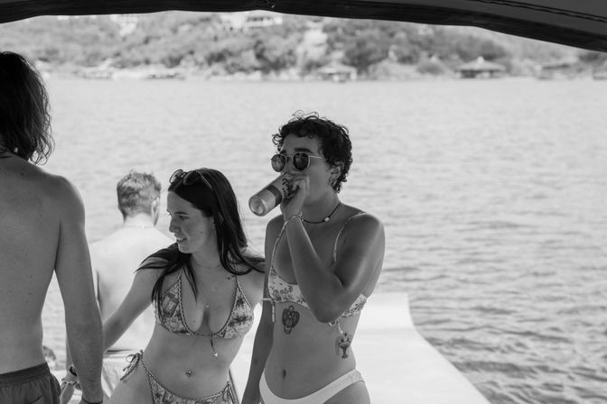 Black-and-white photo of young adults on a boat by a lake, two women in bikinis—one drinking from a can and wearing sunglasses—standing on a dock with water and distant shoreline