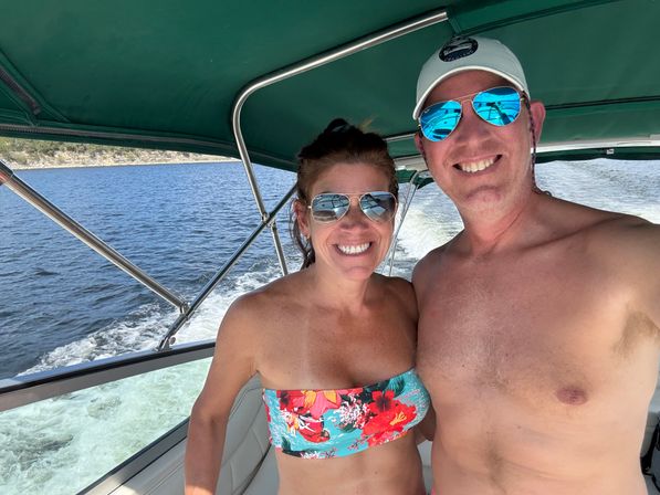 Two smiling adults in mirrored sunglasses on a summer motorboat under a green bimini, woman in a floral bandeau and a shirtless man in a white cap with the boat wake and lakeshore visible behind them
