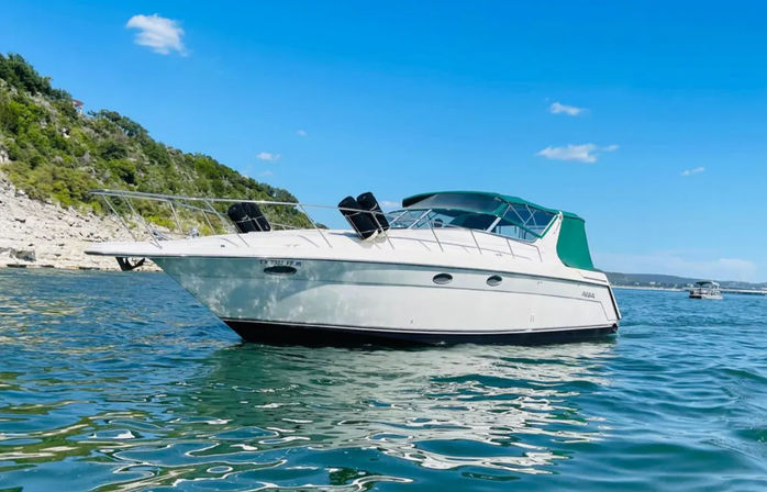 White cabin cruiser motorboat with green canvas top floating on turquoise lake near a rocky, tree-covered shoreline under a bright blue sky