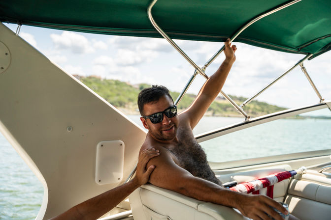 Shirtless man in sunglasses relaxing on a motorboat under a green canopy on a sunny lake, holding a drink with tree-lined shoreline in the background — summer boating scene.