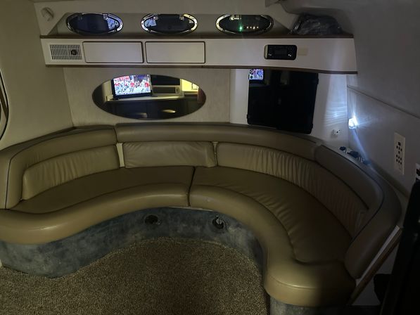 Cozy yacht cabin lounge with curved tan leather banquette, oval porthole windows, overhead storage cabinets, small mirrored TV reflection, and carpeted floor.
